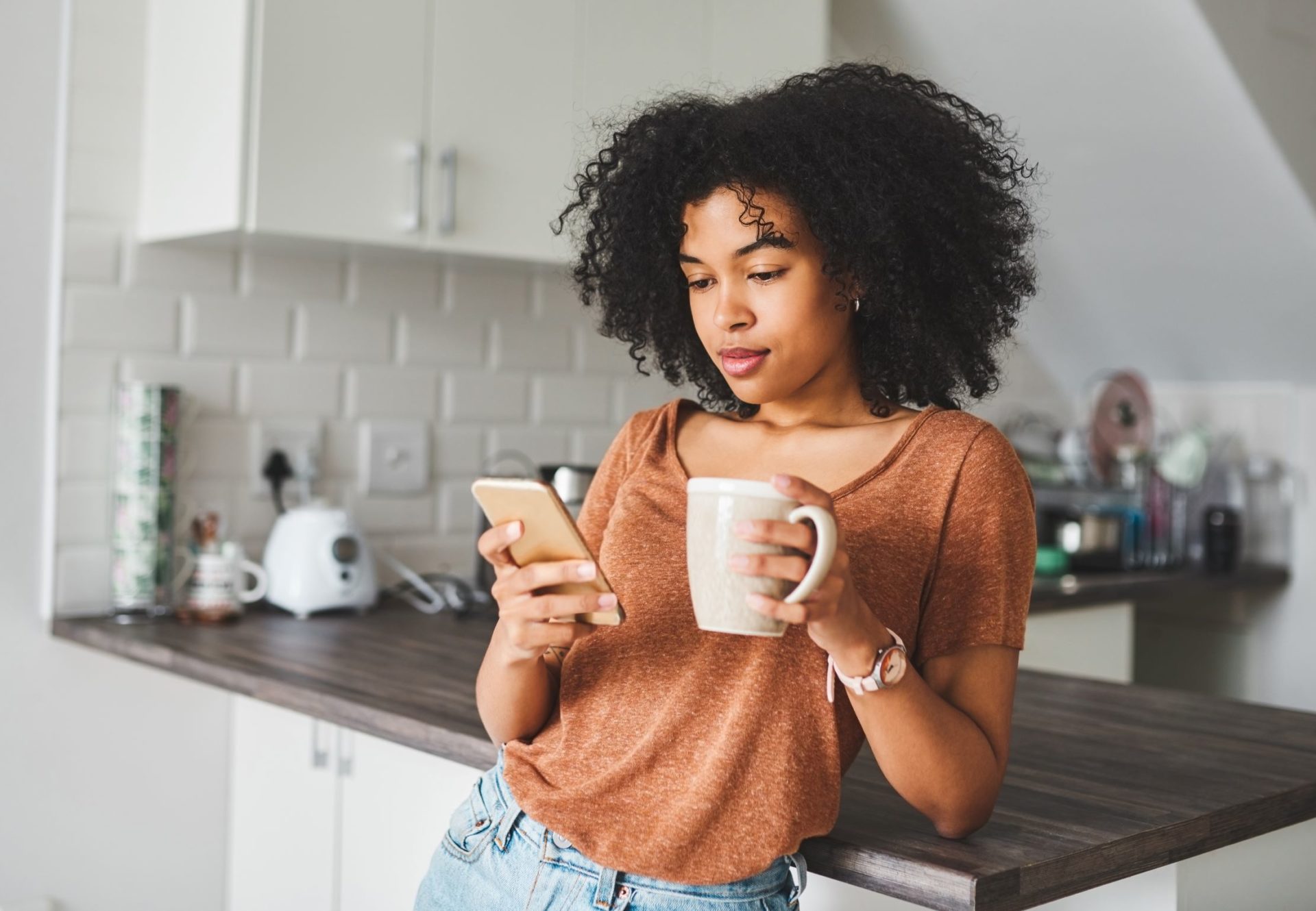 Woman drinking coffee in kitchen with phone in hand